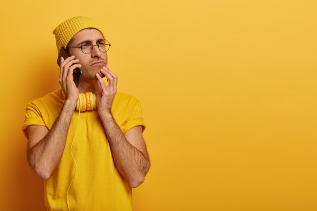 Ernsthafter nachdenklicher junger kaukasischer Mann wendet sich ab, denkt tief über etwas nach, trägt transparente Brille, lässiges gelbes T-Shirt und Kopfbedeckung