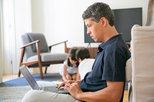 Ernsthafter Profi, der zu Hause arbeitet, auf dem Boden sitzt und einen Laptop benutzt