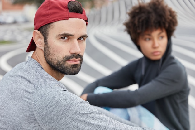 Kostenloses Foto ernsthafter unrasierter mann in stilvoller roter mütze, graues sweathshirt, genießt freizeit, atmet frische luft, afro-mädchen im hintergrund