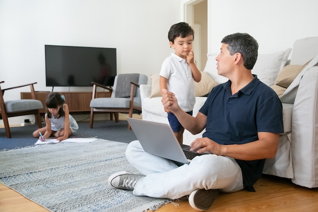 Ernsthafter Vater, der sich um Kinder kümmert, während er zu Hause arbeitet, mit Laptop auf dem Boden sitzt und mit dem kleinen Sohn spricht.