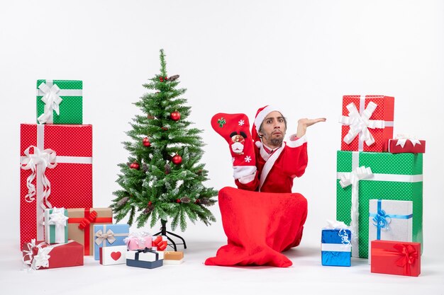 Ernsthafter Weihnachtsmann, der auf dem Boden sitzt und Weihnachtssocke nahe Geschenken und geschmücktem Neujahrsbaum auf weißem Hintergrund trägt