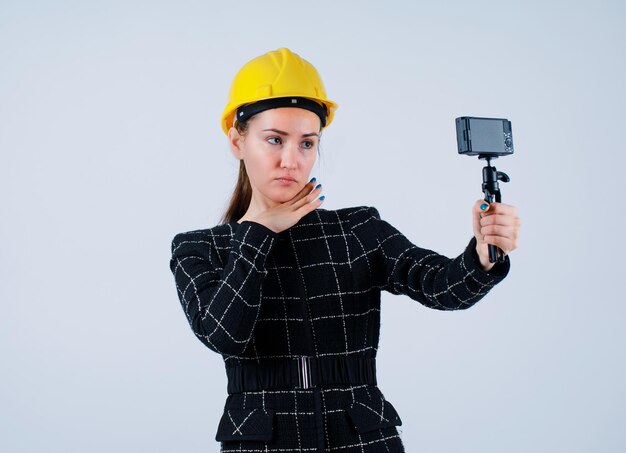 Ernsthaftes Ingenieurmädchen macht Selfie mit ihrer Kamera, indem sie die Hand unter das Kinn auf weißen Hintergrund legt