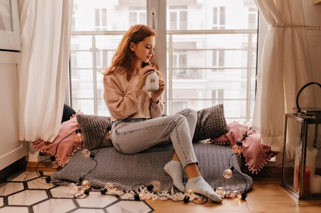 Ernsthaftes Mädchen in lässigen Jeans, das Tee trinkt Innenfoto einer glamourösen Ingwerdame mit einer Tasse Kaffee