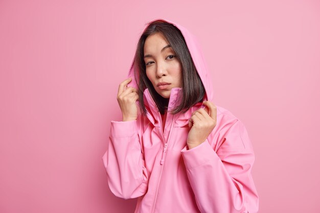 Ernsthaftes selbstbewusstes tausendjähriges Mädchen mit dunklem Haar trägt eine Anorak-Kapuze auf dem Kopf und sieht direkt Modelle gegen die rosa Wand aus, die an einem kalten, windigen Tag spazieren gehen wird Menschen und Stil