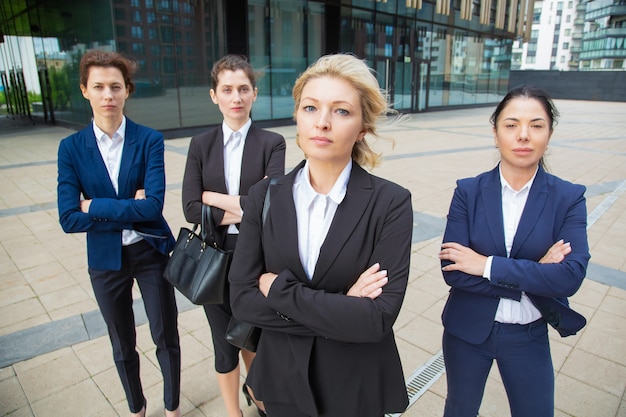 Ernsthaftes selbstbewusstes weibliches professionelles Team, das mit Gruppenleiter zusammen nahe Bürogebäude steht, posiert und Kamera betrachtet. Vorderansicht. Gruppenporträtkonzept der Geschäftsfrauen