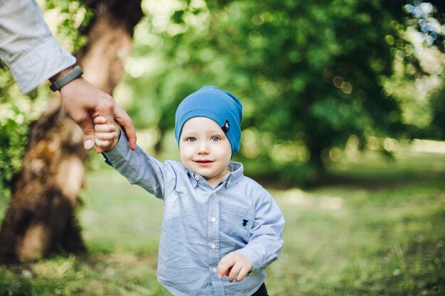 Ernte des Vaters, der den kleinen Jungen, der im Sommerpark spazieren geht, von Hand hält Kleiner Junge in blauer Kappe, der ein kariertes Hemd trägt, lächelt und Spaß draußen in der Natur hat Glückliches Kind und Sommerkonzept