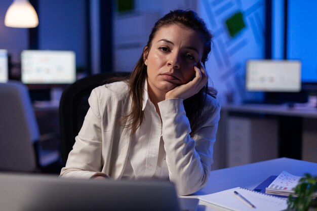 Erschöpfte Geschäftsfrau, die spät in der Nacht im Geschäftsbüro müde in der Kamera schaut und den Kopf in der Handfläche seufzt