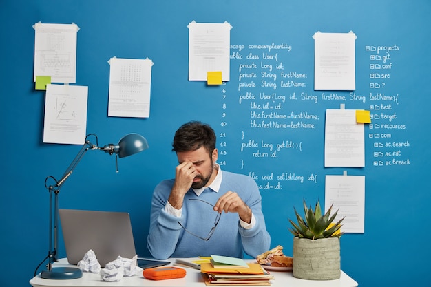 Erschöpfter Geschäftsmann reibt sich die Nase, nimmt die Brille ab, leidet unter Augenbelastung und Kopfschmerzen, hat Probleme bei der Arbeit, sitzt im Coworking Space mit Laptop, blaue Wand mit schriftlichen Notizen.