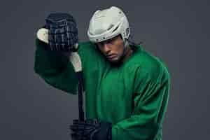 Kostenloses Foto erschöpfter hockeyspieler mit grüner schutzausrüstung und weißem helm, der mit dem hockeyschläger auf grauem hintergrund steht.