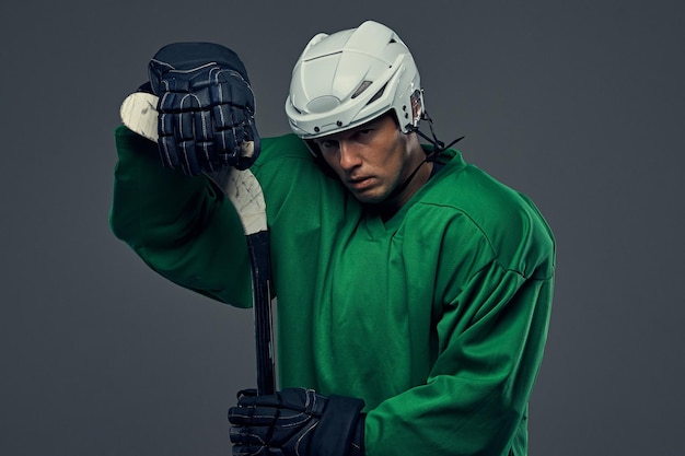 Erschöpfter Hockeyspieler mit grüner Schutzausrüstung und weißem Helm, der mit dem Hockeyschläger auf grauem Hintergrund steht.