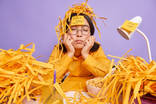 Kostenloses Foto erschöpfter, schläfriger student, der im coworking space überarbeitet ist, arbeitet hart, bevor die frist abgeschlossen ist, hält die hände unter dem kinn, umgeben von papierabfällen, posiert in einer lila wand des kreativen arbeitsplatzes