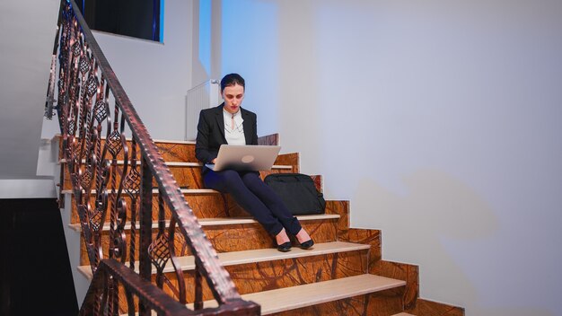 Erschöpfter überarbeiteter Unternehmer, der Überstunden bei der Eingabe der Projektfrist auf dem Laptop macht. Ernster Unternehmer, der an einem Firmenjob arbeitet, der spät in der Nacht auf der Treppe des Geschäftsgebäudes sitzt.