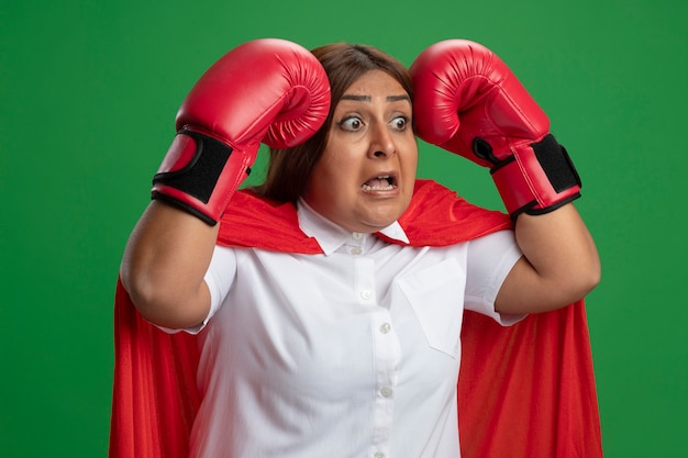 Erschreckte Superheldenfrau mittleren Alters, die Boxhandschuhe hält, die Hände auf Kopf lokalisiert auf grünem Hintergrund halten