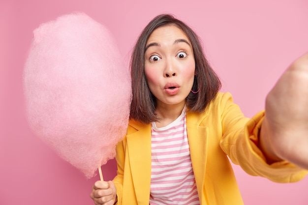 Erschrockene asiatische Frau starrt verwanzte Augen an, macht Foto von sich selbst hält leckere Zuckerwatte, isst gerne etwas Süßes und posiert in stilvoller Kleidung gegen rosa Wand