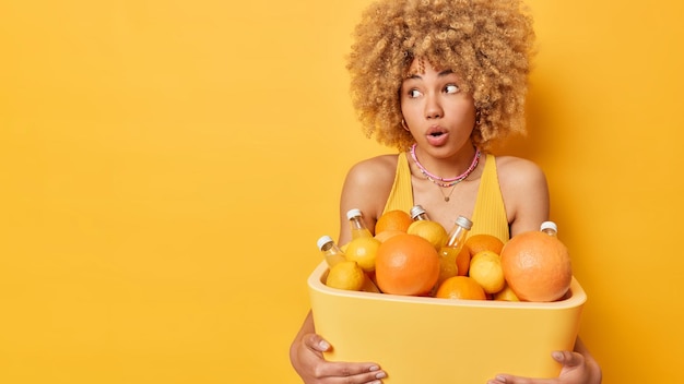 Kostenloses Foto erschrockene hübsche junge frau hält eine tragbare kühlbox mit früchten und getränken starrt beeindruckt weg, gekleidet in ein sommer-t-shirt, das über einem leeren raum auf leuchtend gelbem hintergrund für ihre werbung isoliert ist