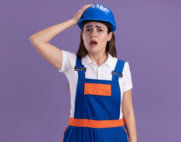 Erschrockene junge Baumeisterin in Uniform, die Hand auf den Kopf legt, isoliert auf lila Wand