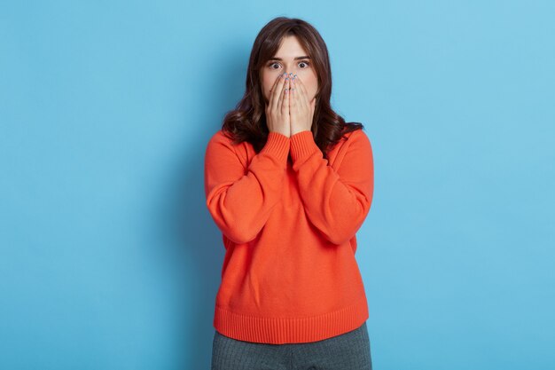 Erschrockene junge erwachsene dunkelhaarige Frau, die lässigen orange Pullover trägt