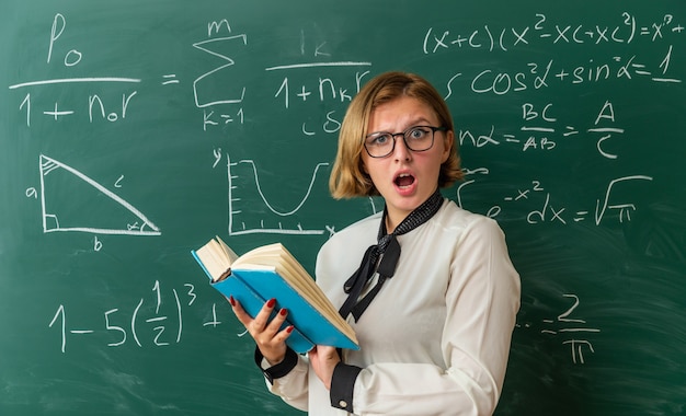 Erschrockene junge Lehrerin mit Brille, die vor der Tafel steht und ein Buch im Klassenzimmer hält