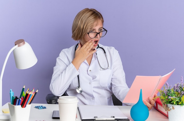 Erschrockene junge Ärztin, die ein medizinisches Gewand mit Stethoskop und Brille trägt, sitzt am Tisch mit medizinischen Instrumenten, die das Notebook auf blauem Hintergrund halten und betrachten