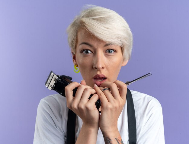 Erschrockene junge schöne Friseurin in Uniform, die eine Schere mit Haarschneidemaschinen auf blauem Hintergrund hält