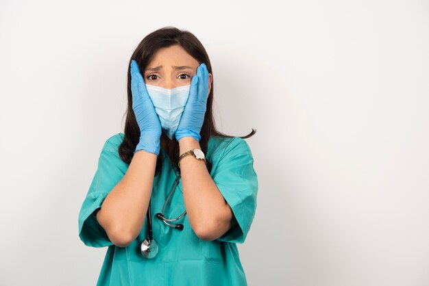 Erschrockener Arzt in der medizinischen Maske und in den Handschuhen, die ihr Gesicht weißen Hintergrund halten.