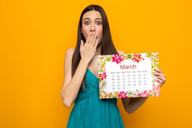 Erschrockener bedeckter Mund mit Hand schönes junges Mädchen am glücklichen Frauentag, der Kalender hält