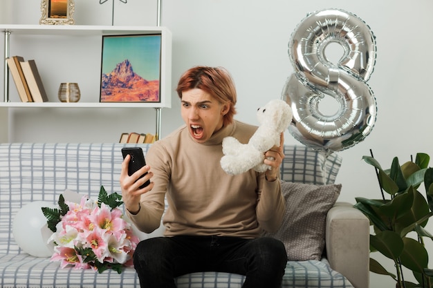 Erschrockener, gutaussehender Kerl am glücklichen Frauentag, der einen Teddybären hält, der auf das Telefon in seiner Hand schaut, das auf dem Sofa im Wohnzimmer sitzt