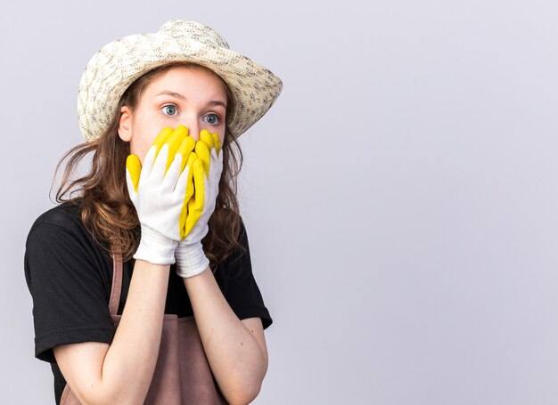 Erschrockener junger Gärtner mit Gartenhut mit Handschuhen bedecktes Gesicht mit Händen isoliert auf weißer Wand mit Kopierraum