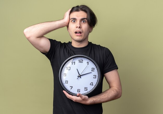 Erschrockener junger gutaussehender Kerl, der ein schwarzes T-Shirt trägt und eine Wanduhr hält, packte den Kopf isoliert auf olivgrüner Wand?