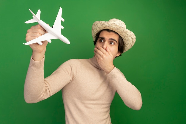 Erschrockener junger gutaussehender Kerl, der Hut trägt und den Mund des Spielzeugflugzeugs mit Hand lokalisiert auf grüner Wand anhebt und betrachtet