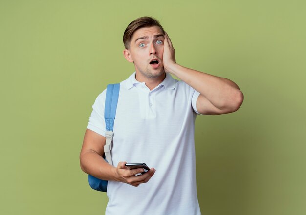 Erschrockener junger hübscher männlicher Student, der Rückentasche hält Telefon hält und Hand auf Wange lokalisiert auf Olivgrün legt