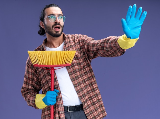 Erschrockener junger hübscher Reinigungsmann, der T-Shirt und Handschuhe hält, die Mopp halten, der Stoppgeste zeigt, die auf blauer Wand lokalisiert wird