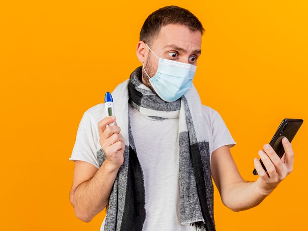 Erschrockener junger kranker Mann, der Wintermütze und medizinische Maske hält, die Thermometer hält und Telefon in seiner Hand lokalisiert auf gelbem Hintergrund betrachtet