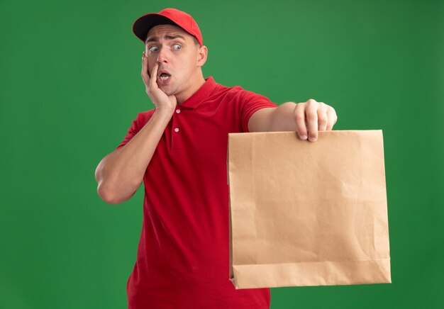 Erschrockener junger Lieferbote, der Uniform und Kappe trägt, die Papiernahrungsmittelpaket an der Kamera hält Hand auf Wange lokalisiert auf grüner Wand hält