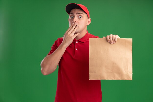 Erschrockener junger Lieferbote, der Uniform und Kappe trägt, die Papiernahrungsmittelpaket bedeckten Mund mit Hand lokalisiert auf grüner Wand hält