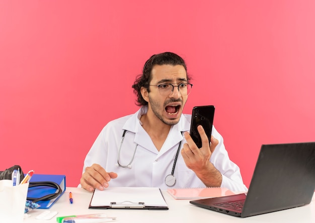 Erschrockener junger männlicher Arzt mit medizinischer Brille, der ein medizinisches Gewand mit Stethoskop trägt, das am Schreibtisch sitzt