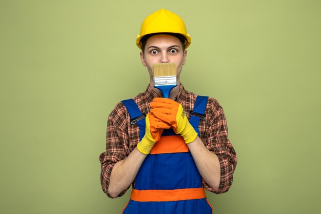Erschrockener junger männlicher Baumeister in Uniform mit Handschuhen und bedecktem Gesicht mit Pinsel