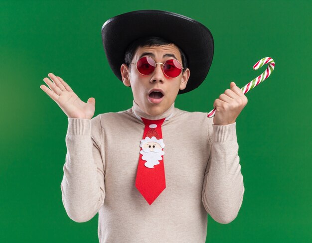 Erschrockener junger Mann, der Hut mit Weihnachtskrawatte und Brille trägt, die Weihnachtsbonbonverbreitungshand lokalisiert auf grüner Wand hält