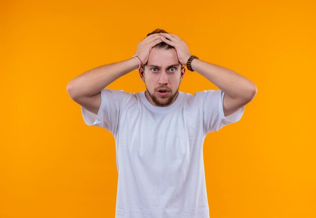 Erschrockener junger Mann, der weißes T-Shirt trägt, ergriff Kopf auf lokalisiertem orange Hintergrund