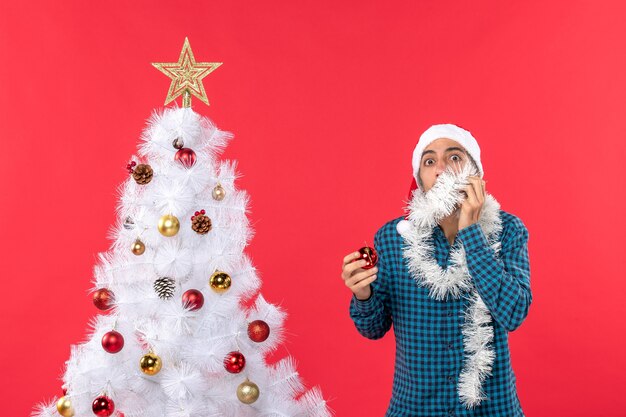 Erschrockener junger Mann mit Weihnachtsmannhut in einem blau gestreiften Hemd und im Dekorationszubehör stehend