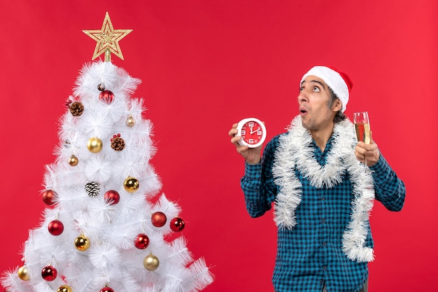Erschrockener junger Mann mit Weihnachtsmannhut und hält ein Glas Wein und eine Uhr, die nahe Weihnachtsbaum auf Rot stehen