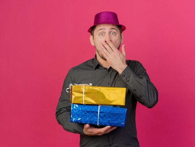 Erschrockener junger Party-Typ, der rosa Hut trägt, der Geschenkboxen bedeckte, bedeckte Mund mit Hand lokalisiert auf Rosa