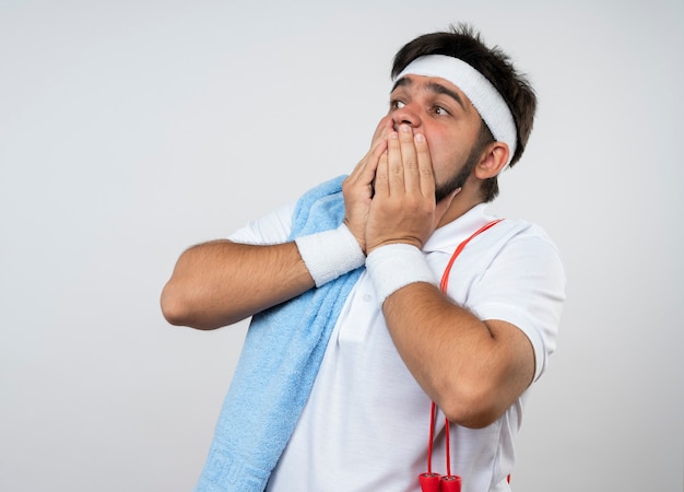 Erschrockener junger sportlicher Mann, der seitliches Tragen von Stirnband und Armband mit Handtuch und Springseil auf schulterbedecktem Mund mit auf weißer Wand isolierten Händen betrachtet