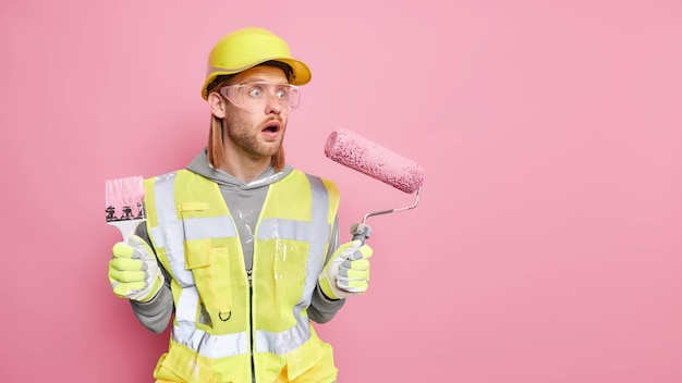 Kostenloses Foto erschrockener männlicher industriearbeiter macht reparaturen in der wohnung hält malerrolle und pinsel starrt mit schockiertem ausdruck weg