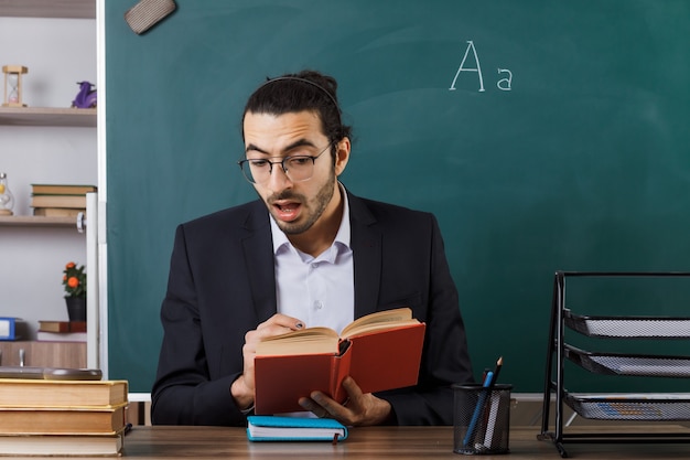 Erschrockener männlicher Lehrer mit Brille, der ein Buch hält und liest, das am Tisch mit Schulwerkzeugen im Klassenzimmer sitzt