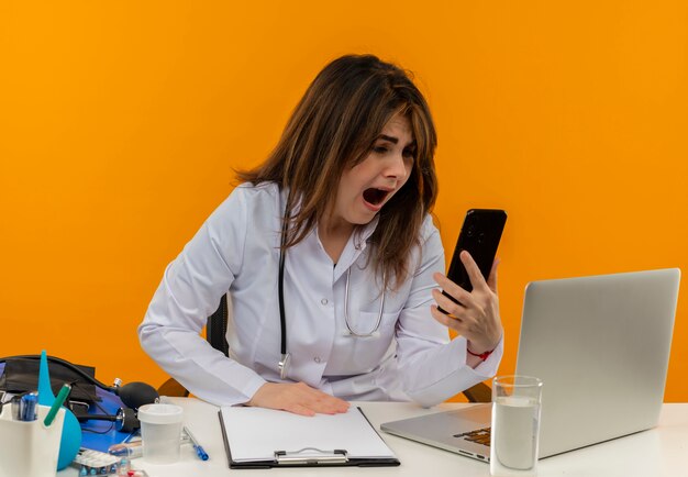 Erschrockener weiblicher Arzt mittleren Alters, der medizinische Robe mit Stethoskop trägt, das am Schreibtisch sitzt, arbeitet am Laptop mit medizinischen Werkzeugen, die Telefon auf lokalisiertem orangefarbenem Hintergrund mit Kopienraum halten und betrachten