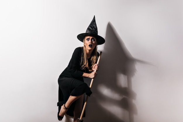 Erschrockener weiblicher Zauberer, der magischen Besen hält. Innenfoto der verängstigten Frau im Hexenkostüm, das in Halloween aufwirft.