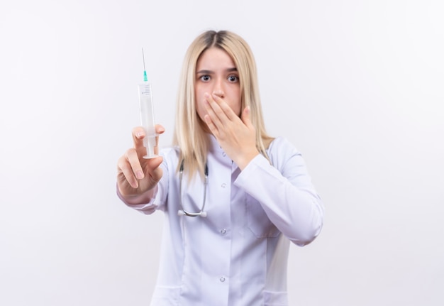 Erschrockenes junges blondes Mädchen des Arztes, das Stethoskop und medizinisches Kleid hält Spritzen bedeckte Mund auf lokalisiertem weißem Hintergrund