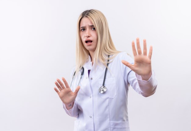 Erschrockenes junges blondes Mädchen des Arztes, das Stethoskop und medizinisches Kleid trägt, das Stoppgeste mit beiden Händen auf lokalisiertem weißem Hintergrund zeigt