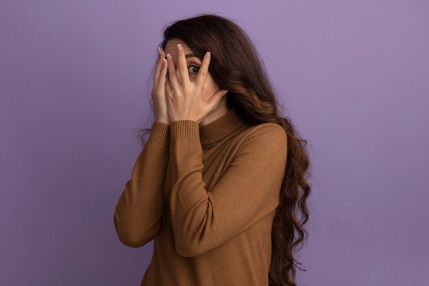 Erschrockenes junges schönes Mädchen, das braunes Rollkragenpullover-bedecktes Gesicht mit den Händen auf lila Wand isoliert trägt